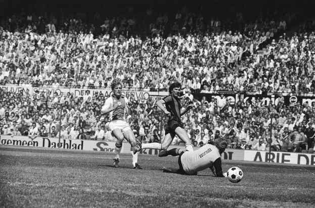 Schwarzes Foto von Männern, die auf einem Feld Fußball spielen, mit Zuschauern im Hintergrund und einem Banner mit der Aufschrift 'Bundesliga 1978-79 Ajax 1-1 Hannover 96'.