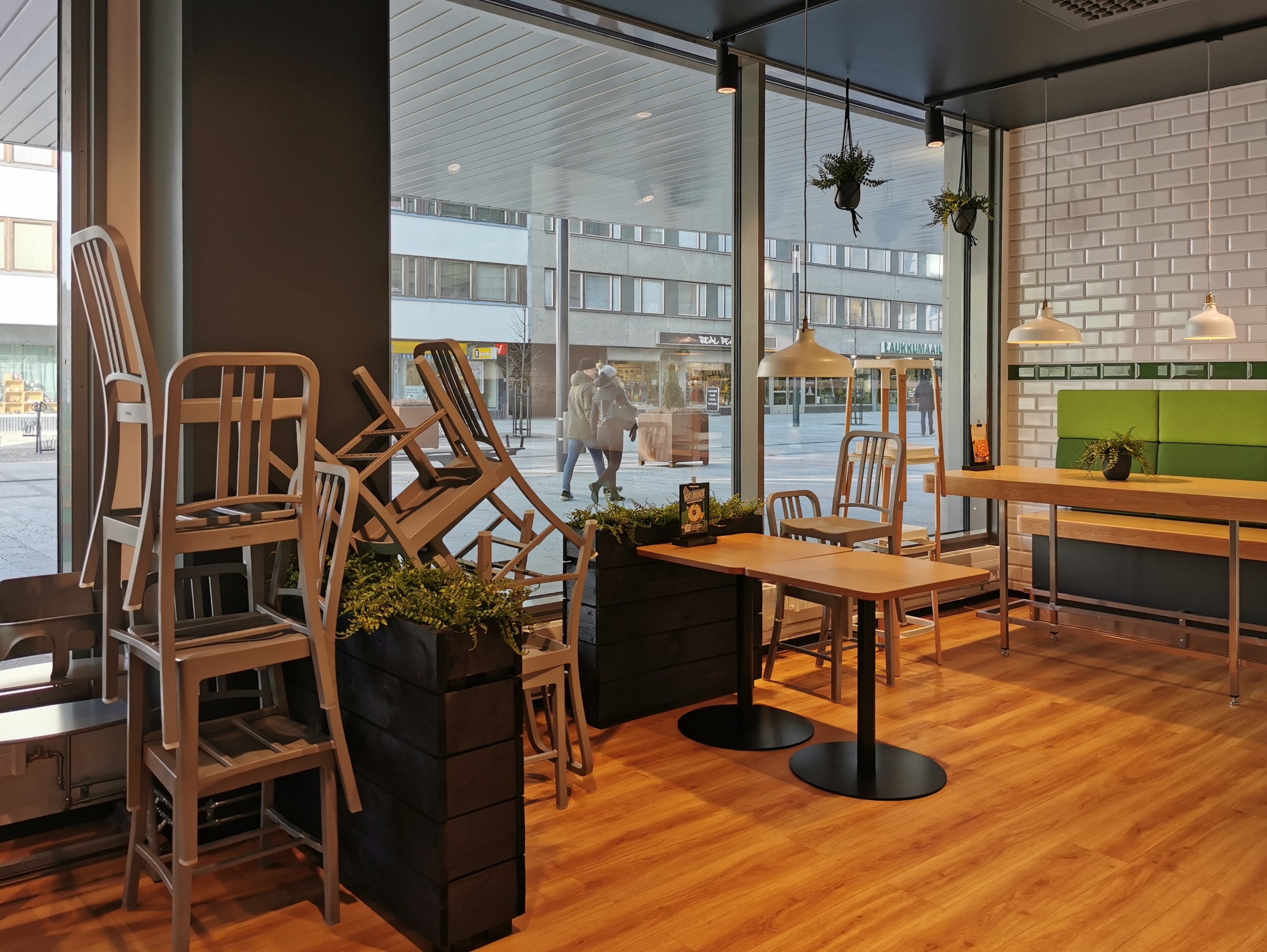 Ein Restaurant-Innenraum mit hölzernen Tischen und Stühlen vor einem großen Fenster, das eine Berliner Straßenszene mit Fußgängern, Gebäuden und städtischen Elementen zeigt.