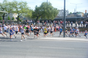Gruppe von Läufern bei einem Marathon an einer Ziellinie mit einer Schleife vorbei, umgeben von einer Absperrung, Barrieren, Pfosten, Schildern, einer Brücke, Gebäuden, Bäumen und einem bewölkten Himmel.