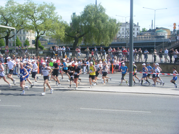 Gruppe von Läufern bei einem Marathon an einer Ziellinie mit einer Schleife vorbei, umgeben von einer Absperrung, Barrieren, Pfosten, Schildern, einer Brücke, Gebäuden, Bäumen und einem bewölkten Himmel.