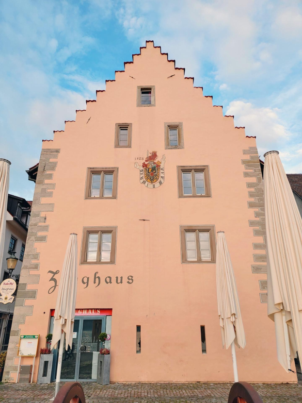 Ein rosafarbenes Gebäude mit einem zentralen Uhrenturm, umgeben von Fenstern, Schirmen, einem Schild, einer Straßenlaterne, Topfpflanzen und einem bewölkten Himmel, repräsentiert München, Deutschland.