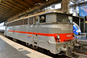 Eine Lokomotive auf den Schienen im Vordergrund mit einer Plattform links und einem Schuppen oben; im Hintergrund ein weiterer Zug, eine Plattform, ein Banner und Pfosten.