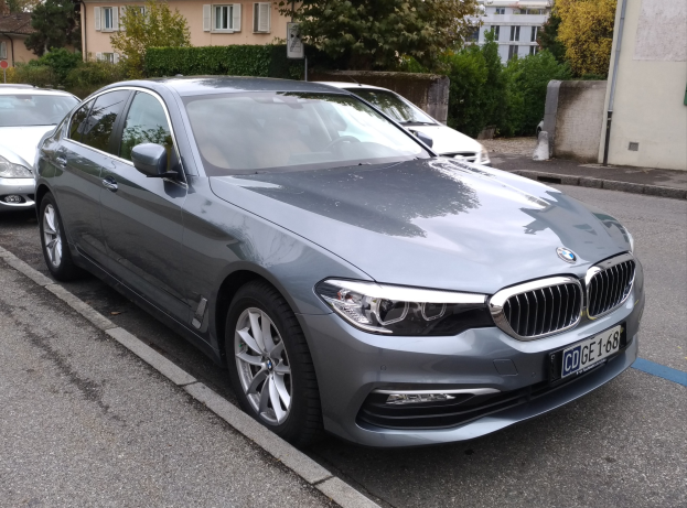 Ein BMW 5er 520d xDrive Luxury Line fährt auf einer Straße mit Gebäuden, Bäumen und einem klaren blauen Himmel im Hintergrund.