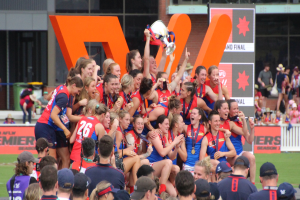 Gruppe von Frauen-AFL-Spielerinnen feiert mit einem Pokal vor einem Publikum, trägt Sportuniformen und hält den Pokal in den Händen.