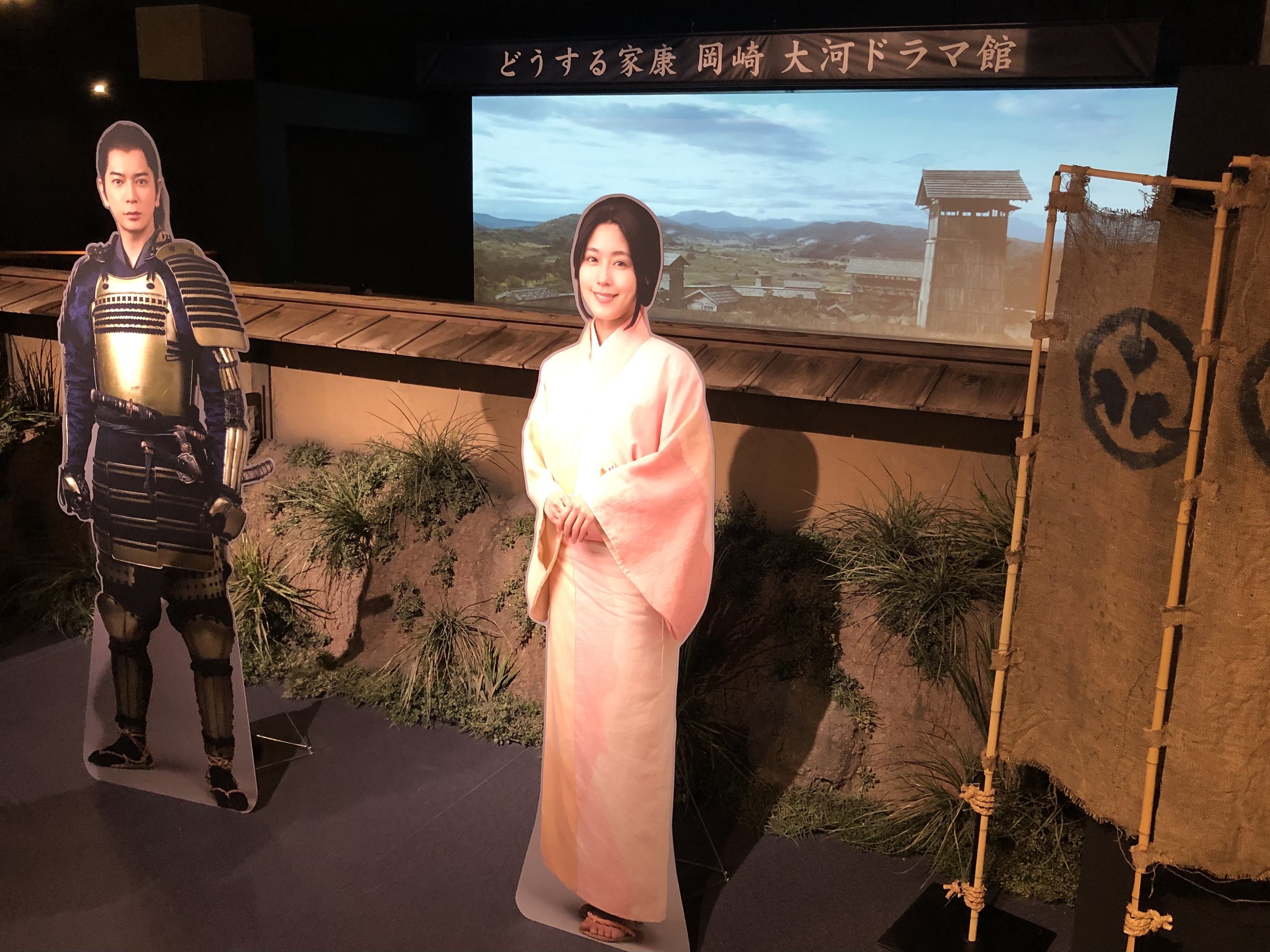 Zwei Menschen in traditioneller japanischer Kleidung - eine Frau in einem pinken Kimono und ein Mann in Samurai-Rüstung - lächeln gemeinsam in einer Szene aus einem japanischen Film, mit Pflanzen, einem Bildschirm und Lichtern im Hintergrund.
