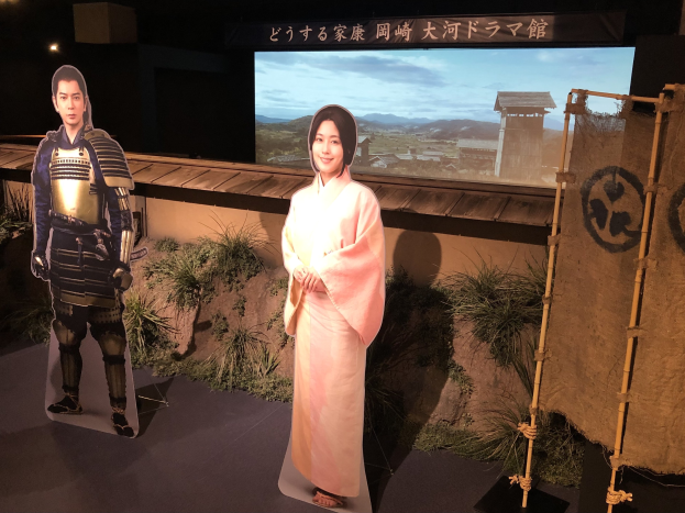 Zwei Menschen in traditioneller japanischer Kleidung - eine Frau in einem pinken Kimono und ein Mann in Samurai-Rüstung - lächeln gemeinsam in einer Szene aus einem japanischen Film, mit Pflanzen, einem Bildschirm und Lichtern im Hintergrund.