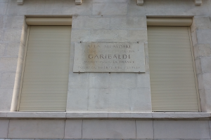 Ein zweistöckiges Gebäude mit zwei Fenstern und Läden sowie einer Tafel an der Seite, wahrscheinlich ein Denkmal.