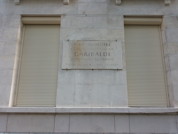 Ein zweistöckiges Gebäude mit zwei Fenstern und Läden sowie einer Tafel an der Seite, wahrscheinlich ein Denkmal.