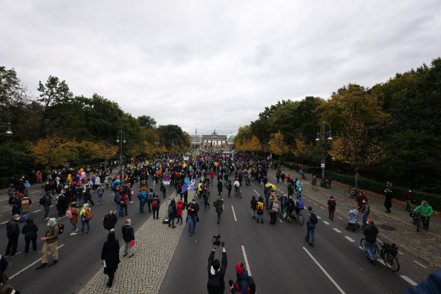 Eine große Gruppe von Menschen marschiert auf einer Straße mit Bäumen und Laternen in Berlin, einige halten Kameras, mit einem Gebäude und einem klaren Himmel im Hintergrund.