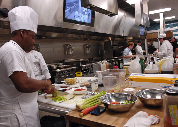Eine Gruppe von Köchen in einer Küche, mit einem Koch im Vordergrund in einem weißen Kleid und einer Mütze, der ein Messer hält und von verschiedenen Kochutensilien, Schüsseln, Gemüse, einem Mobiltelefon und Flaschen umgeben ist, während im Hintergrund ein Fernseher und Deckenleuchten zu sehen sind.