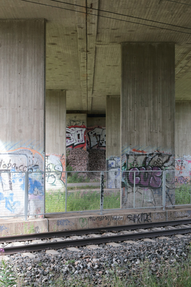Eisenbahnschiene mit Graffiti an einer Betonwand, umgeben von Steinen, Gras, einem Zaun, Stützen, Oberleitungen und einer Dachkonstruktion.