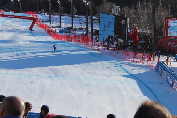 Gruppe von Zuschauern auf einer schneebedeckten Anhöhe, die einen Skifahrer beim Abfahren beobachten, mit Bannern, Absperrungen, Beleuchtung, Bäumen und einem Bildschirm im Hintergrund.