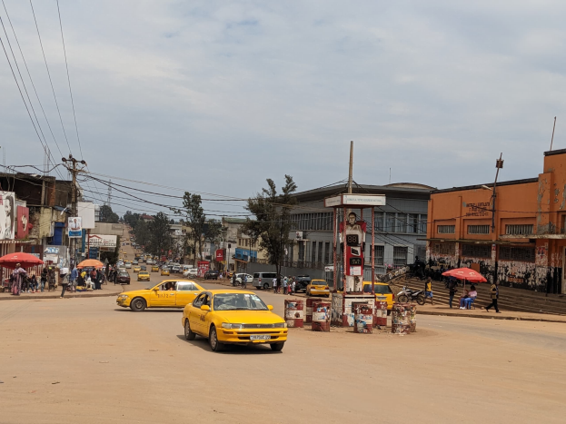 Ein gelbes Taxi fährt eine belebte Stadtstraße mit Gebäuden an beiden Seiten hinunter, während Fußgänger mit Schirmen auf dem Gehweg unter einem bewölkten Himmel gehen, mit Strommasten und Leitungen im Hintergrund.