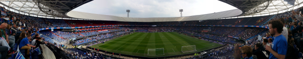 Großes Stadion voller Zuschauer bei einem Fußballspiel, einige halten Kameras, mit einem Netz auf dem Boden und Laternen im Hintergrund unter einem klaren blauen Himmel.
