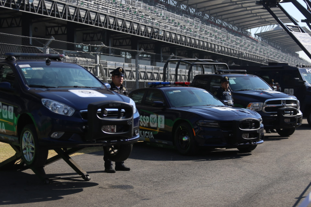 Eine Gruppe von Polizeiwagen vor einer Rennstrecke geparkt, uniformierte Personen auf der Straße stehend und ein Stadion mit Geländern und Maschendrahtzaun im Hintergrund.