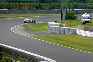 Ein Rennauto fährt eine Rennstrecke hinunter mit einem Polizeiwagen im Hintergrund, umgeben von Gras, Pflanzen, Bäumen und Zaun, mit einem Fahrzeug auf der rechten Seite und Text unten.