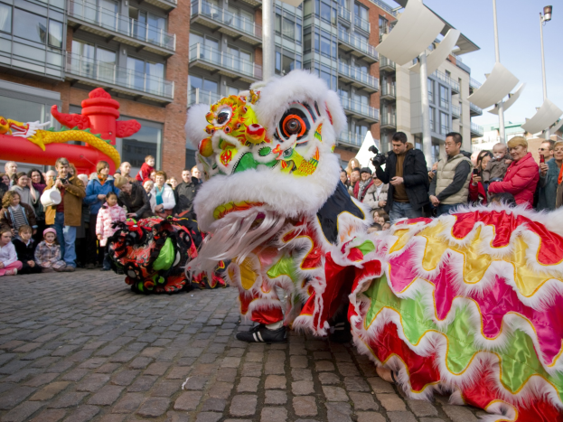 Ein farbenfrohes chinesisches Neujahrsfest in Amsterdam mit einer Löwen-Tanzvorstellung vor einer Zuschauermenge, einige halten Kameras, vor einer Kulisse aus Gebäuden, Laternenmasten und einem klaren blauen Himmel.