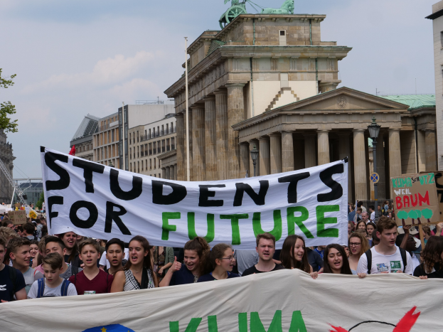 Gruppe von Schülern marschiert in Berlin mit einer bunten "Students for Future"-Schriftzug vor dem Hintergrund von Gebäuden, Bäumen und Himmel.
