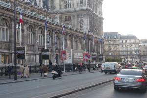 Eine belebte Stadtstraße mit Fahrzeugen, Fußgängern, hohen Gebäuden, Laternen, Flaggen, Texttafeln und einem Geländer unter einem sichtbaren Himmel.
