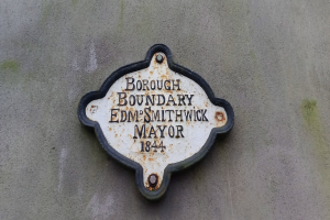 Metallschild an der Seite eines Gebäudes, das die Borough-Grenze markiert, mit schwarzem Hintergrund und weißer, fettgedruckter, zentrierter Schrift, die "Borough Boundary Edm Smithwick Mayor" lautet.