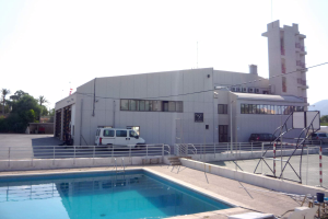 Ein großes Gebäude mit einem Swimmingpool davor, umgeben von einem Metallzaun, Fahrzeugen, Pfählen, Drähten, einem Straßenmast, Bäumen, Hügeln und einem bewölkten Himmel.