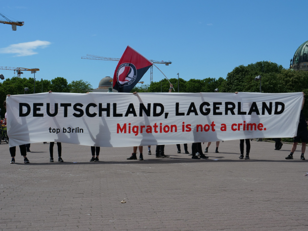 Gruppe von Menschen mit einem Banner 'Deutschland, Lagerland Migration ist kein Verbrechen' und einer Flagge, im Hintergrund Straßenmasten, Straßenlaternen, Bäume, Kräne, ein Gebäude und ein bewölktes Himmel.