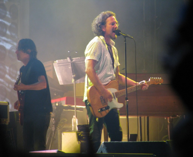 Live-Musikauftritt auf der Buhne mit einem Mann in weissem Hemd, der in ein Mikrofon singt und Gitarre spielt, begleitet von einem anderen Mann in schwarzem Hemd, der Gitarre spielt, mit Piano, Kabeln, Lautsprechern und Buhnenlicht im Hintergrund.