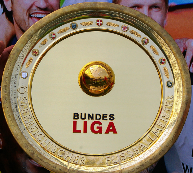 Mann hält goldenen Schild mit "Bundesliga"-Text hoch, im Hintergrund eine Fahne mit drei Personen