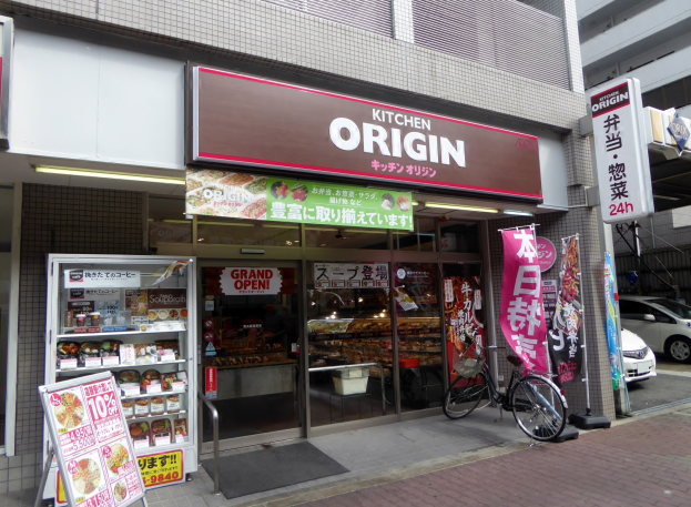 Ladenfront mit Glaswänden und -türen, mit einem parkenden Fahrrad davor, Lebensmitteln im Inneren und Texttafeln; Fahrzeuge auf der benachbarten Straße sichtbar.
