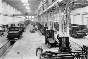 Ein Schwarz-Weiß-Foto einer Fabrikhalle mit Arbeitern, die an Zügen auf dem Boden arbeiten, umgeben von Metallstangen, Deckenlampen und Fenstern an den Wänden.