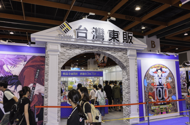 Menschen vor einem Spielshow-Stand auf einer Messe, mit Werbetafeln und Deckenbeleuchtung.