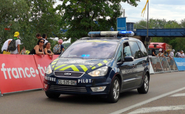 Ein Polizeiwagen fährt an einer Menschenmenge vorbei, die Schilder hölt, eine Brücke, Bäume, eine Fahne auf einem Mast und einen bewölkten Himmel.