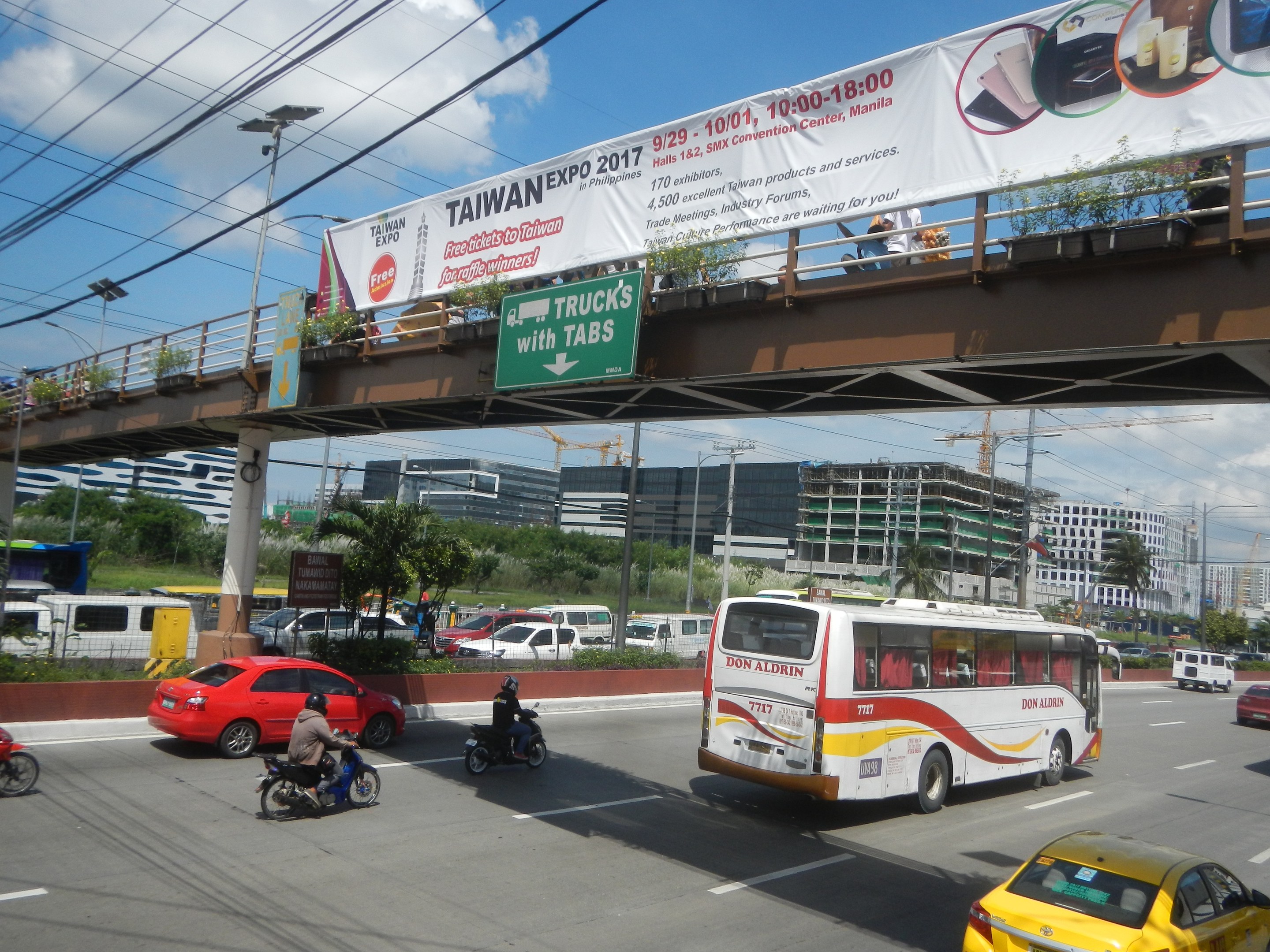 Eine belebte Stadtstraße mit Autos, Bussen, Motorrädern, einer Brücke mit Geländern und einem Banner, Laternenmasten, Strommasten mit Drähten, Bäumen, Gebäuden und einem bewölkten Himmel im Hintergrund.