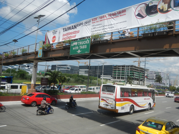 Eine belebte Stadtstraße mit Autos, Bussen, Motorrädern, einer Brücke mit Geländern und einem Banner, Laternenmasten, Strommasten mit Drähten, Bäumen, Gebäuden und einem bewölkten Himmel im Hintergrund.