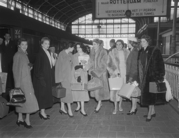 Schwarz-weißes Bild von Frauen, die an einem Bahnhof stehen, mit einem Zug auf der linken Seite und Geländern und einer Tafel auf der rechten Seite, jede hält eine Tasche.