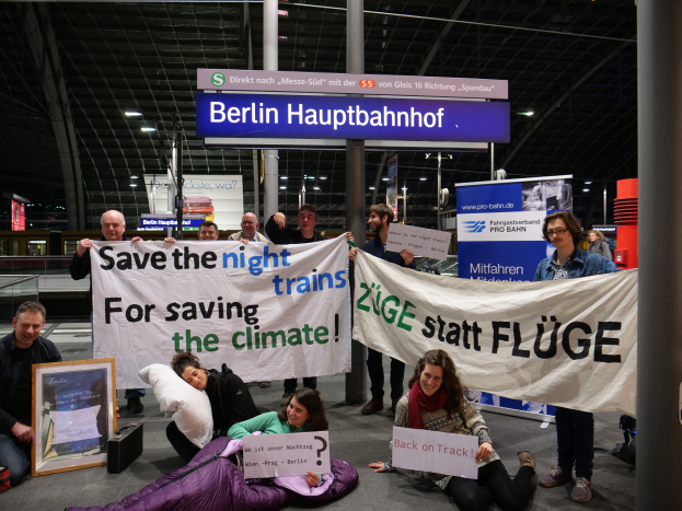 Eine Gruppe von Menschen sitzt auf dem Boden des Berliner Hauptbahnhofs, hält ein Banner mit der Aufschrift "Retten Sie die Nachtzüge zum Schutz des Klimas", umgeben von Kissen, einem Foto Rahmen und anderen Gegenständen, sowie Tafeln mit Text, Säulen, Deckenleuchten und Geländern im Hintergrund.