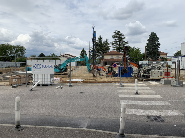 Baustelle mit Geräten einschließlich Pfosten, einem Zaun, einer Tafel, Fahrzeugen, Bäumen, Häusern, Drähten und einem bewölkten Himmel.
