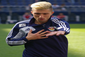 Ein junger Mann in einem blauen T-Shirt steht auf einem Fußballfeld und streckt die Arme aus, zeigt einen entschlossenen Gesichtsausdruck und hat einen unscharfen Hintergrund.