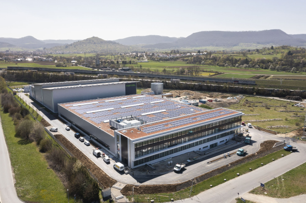 Luftaufnahme eines großen Gebäudes mit Solarmodulen auf dem Dach, umgeben von Fahrzeugen, Bäumen, Gras und Polen, mit Hügeln und einem klaren blauen Himmel im Hintergrund; das Gebäude ist die erste solarbetriebene Fabrik Deutschlands.