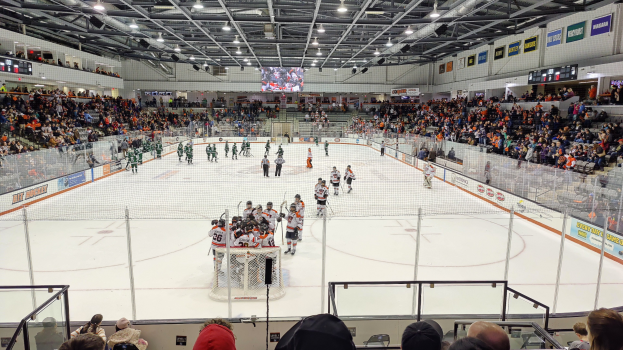 Eishockeyspiel in einer großen Arena mit Spielern auf dem Eis, einem Tor, Zuschauern in den Rängen, Deckenbeleuchtung und Bildschirm sowie Bannern darüber.