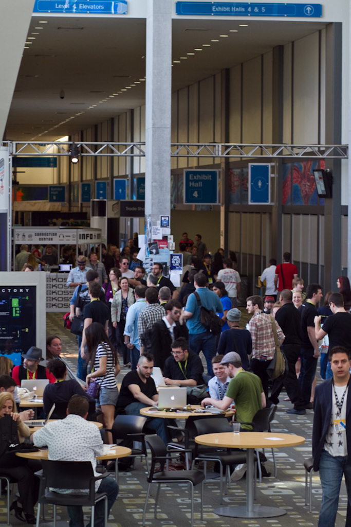 Ein belebter Flughafenvorraum mit wartenden Menschen, einige stehen und andere arbeiten, Richtungsschilder an der rechten Wand und darüber sowie von der Decke hängende Lampen.