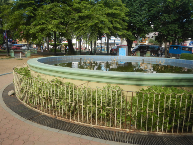 Ein parküberdeckter Brunnen mit Metallzaun umgeben von B├Ąumen, Pflanzen, einem Schild, Fahrzeugen, Menschen, Masten mit Flaggen, Geb├Ąuden mit Fenstern und einem bew├Âlktem Himmel.