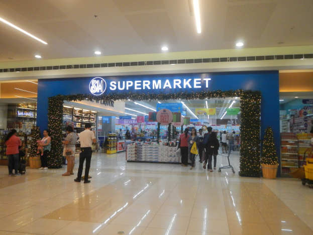 Eine Gruppe von Menschen steht vor einem Supermarkt, einige halten Einkaufstäscher, mit einem Einkaufswagen, einem Topf mit Pflanze, Regalen mit Gegenständen, Schildern und einer Decke mit Leuchten.