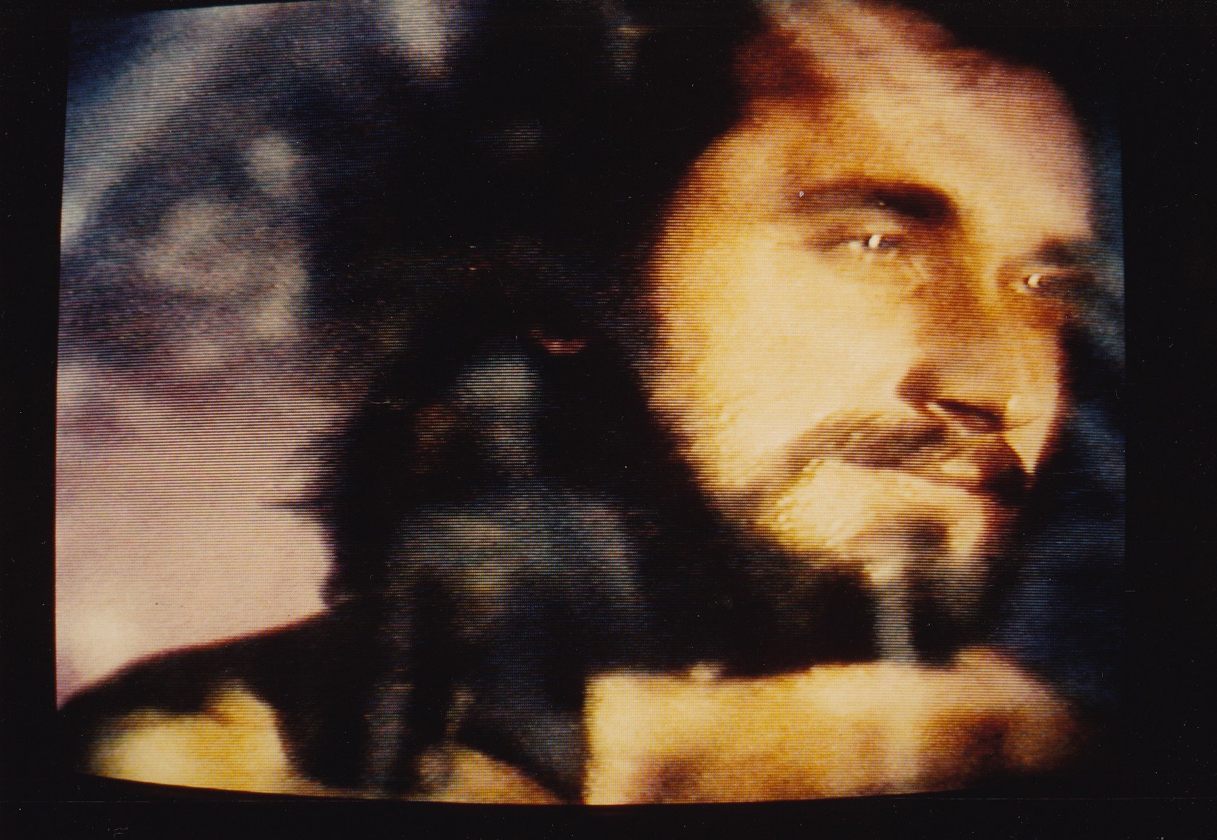 Ein Mann mit langem dunklen Bart und Haar, der ein weißes Hemd trägt, ist auf einem Fernseher mit dunklem Hintergrund zu sehen, sein ernster Blick ist auf den Betrachter gerichtet.