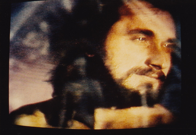 Ein Mann mit langem dunklen Bart und Haar, der ein weißes Hemd trägt, ist auf einem Fernseher mit dunklem Hintergrund zu sehen, sein ernster Blick ist auf den Betrachter gerichtet.