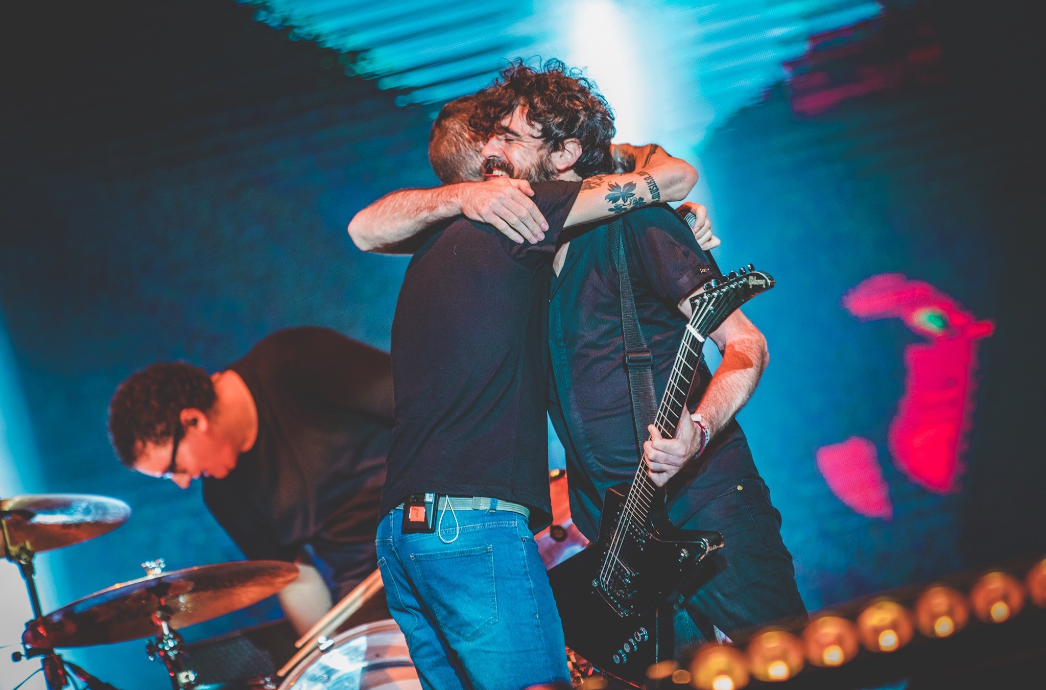 Zwei Männer umarmen sich auf der Bühne eines Konzerts, wobei einer eine Gitarre spielt und ein anderer am Schlagzeug sitzt, unter Bühnenbeleuchtung.