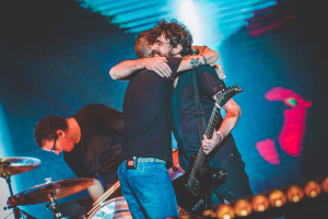 Zwei Männer umarmen sich auf der Bühne eines Konzerts, wobei einer eine Gitarre spielt und ein anderer am Schlagzeug sitzt, unter Bühnenbeleuchtung.