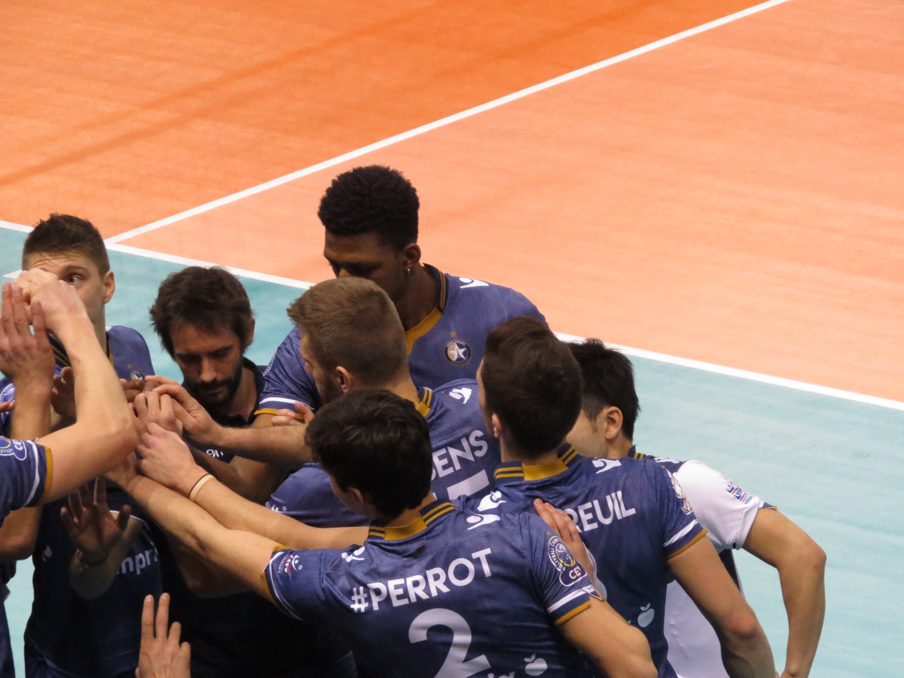 Eine Gruppe von Männern in blauen "Ligue 1"-T-Shirts, die auf einem Volleyballfeld stehen und triumphierend die Hände heben.