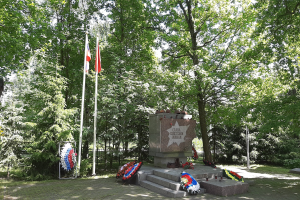 Denkmal in einem Park mit Flaggen, umgeben von Bäumen unter einem klaren Himmel, Blumensträuße auf Stufen und ein Gebäude links.