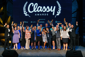 Gruppe von Menschen auf einer Bühne, die Auszeichnungen halten, mit einem Bildschirm im Hintergrund, auf dem "Classy Awards 2019" angezeigt wird.